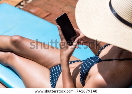 Young Beautiful lady wearing bikini using mobile phone sitting on chair in swimming pool blue water