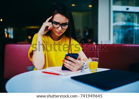 Tired female student in spectacles watching boring training webinar on smartphone connected to wireless internet.Young hipster girl using online banking application on modern smartphone device