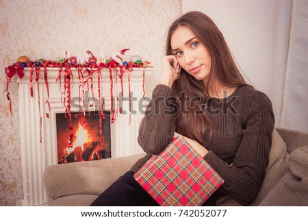 Happy brunette woman holding christmas gift with red tinsel sitting on couch near fireplace at home