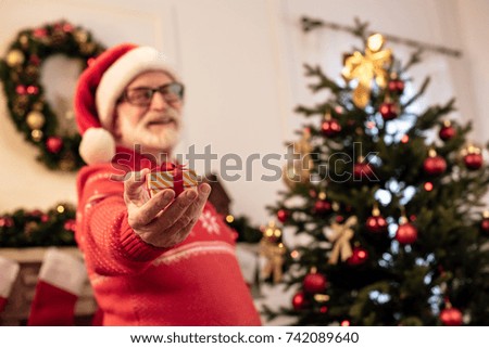 selective focus of senior man in santa hat holding little gift and standing at christmas tree  