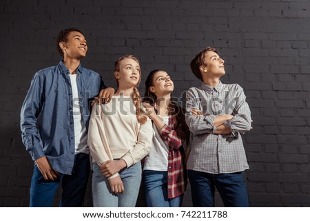 group of stylish teenagers standing together in front of black brick wall and looking away