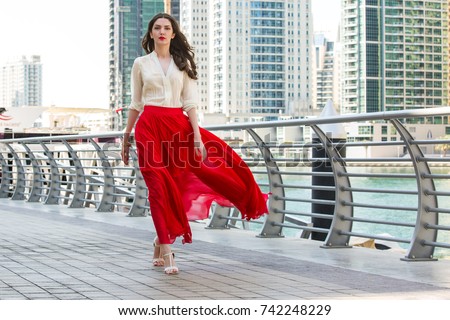 Attractive, Slim, Beautiful And Brunette Girl Wearing White Shirt And Maxi Long Red Silk Fabric Skirt Fly And Wave In The Wind Walking Alongside Dubai Marina Middle East Fashion Concept Royalty-Free Stock Photo #742248229