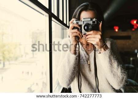 Woman in white sweater stands with camera before a bright window