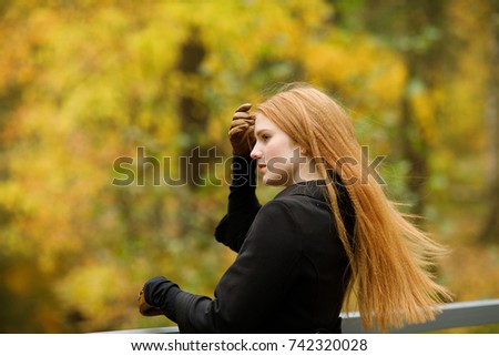 Portrait of a stylish young red-haired woman in a black coat styling  hair against the background of autumn trees for a walk in the park. Human facial expressions, emotions and feelings.
