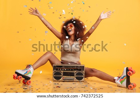 Charming young african woman in retro wear and roller scates throwing confetti while sitting with boombox, isolated on yellow background