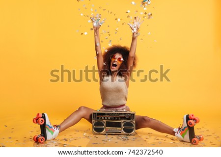 Emotional african disco woman in retro wear and roller scates throwing confetti while sitting with boombox, isolated on yellow background Royalty-Free Stock Photo #742372540