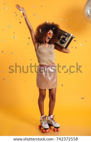 Full length portrait of beautiful overjoyed afro american disco woman with raised hand, standing on roller skates, holding boombox, isolated on yellow background Royalty-Free Stock Photo #742372558