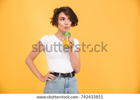 Portrait of a cute casual girl drinking orange juice from a glass and looking at camera isolated over yellow background Royalty-Free Stock Photo #742433851