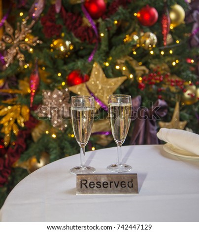 Celebrating Christmas and the New Year with two filled champagne glasses in front of a Christmas Tree covered in baubles on a white table cloth with a reserved sign in front