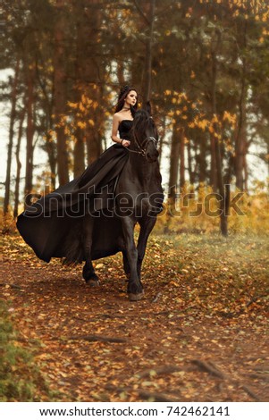 A young girl witch a black widow in a black dress and a tiara gallops horseback on a Friesian horse in the morning fog