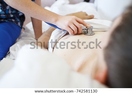 The doctor listens to the patient with a stethoscope who lies in bed with white sheets