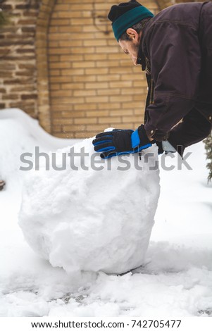man in a jacket and winter gloves rolls a large snowball in the yard of a private house
