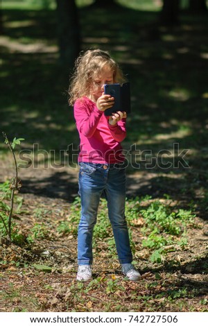Little Cute Girl with Blond Hair is Using Blue Smartphone Standing in the Park. Female Child Taking Photo by Mobile Phone Outside.