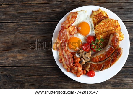 full english breakfast - bean, hash brown, fried eggs sunny side up, bacon slices, sausages, tomatoes, mushrooms on white plate on dark wooden table, view from above  Royalty-Free Stock Photo #742818547