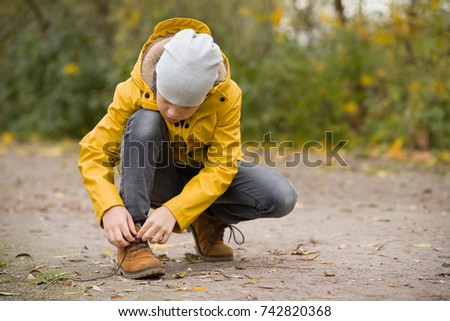 Portrait of cute kid boy in yellow jacket tying laces while walking in the park on a cold autumn day.