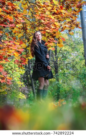 Young girl in classic black coat and blue boots walks in the Park . Autumn Park , green , red and yellow leaves lie on the ground. The girl has long black hair and slim figure .