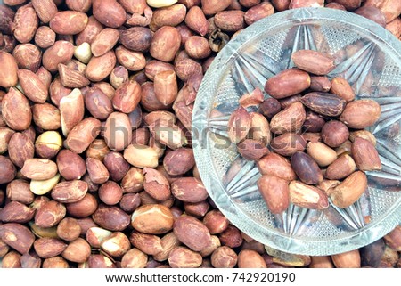 Some fried peanuts in small glass bowl and lot of roasted peanut fruits as background  top view closeup