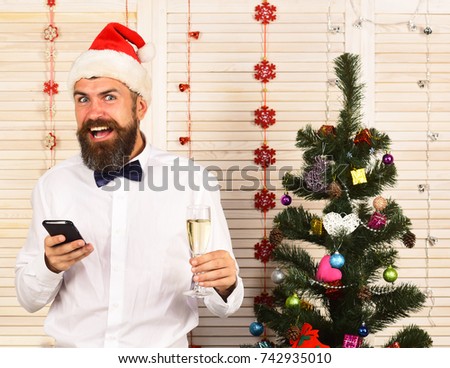Guy near Christmas tree on wooden wall background. Man with beard and bow tie holds champagne glass and phone. Santa Claus in red hat with cheerful face. Celebration and New Year greetings concept