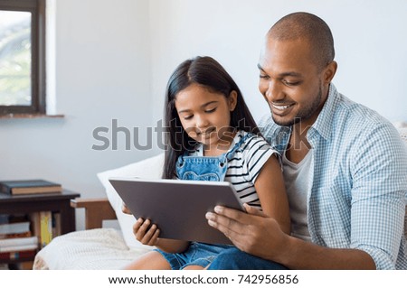 African father and smiling daughter sitting on sofa using digital tablet. Happy dad watching cartoon with little girl. Cheerful father and daughter sitting on couch at home and playing with computer.