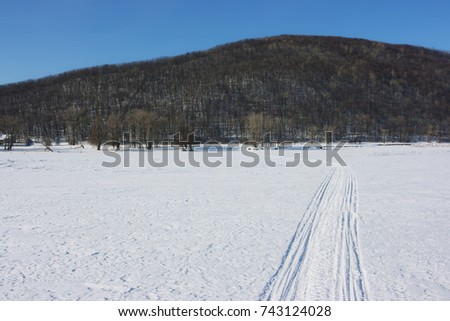 Snowmobile track marks on the snow of a frozen river against mountains and a blue sky