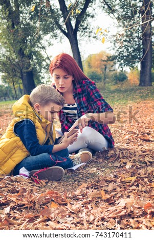 Small kid and his mother sitting in autumn leaves and watching cartoons online. Young mother teaching son how to use mobile phone in the park.