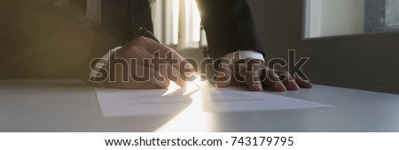 Panorama view of businessman in a shadowy office signing contract or document with a bright light flare from behind.