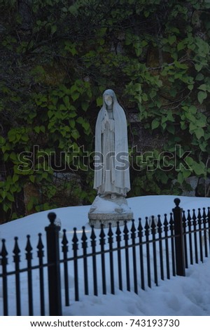 Virgin mary sculpture standing over a mountain of white cold ice snow behind a fence with leaves bushes background 