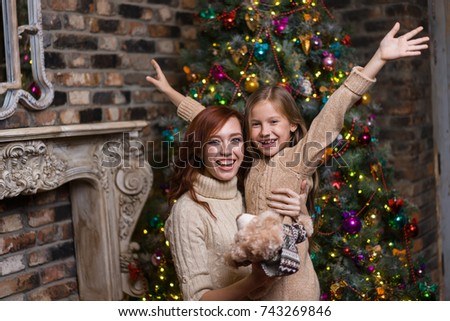 Young attractive woman and her little sweet daughter giving each other presents, hugging and laughing at the background of a Christmas tree