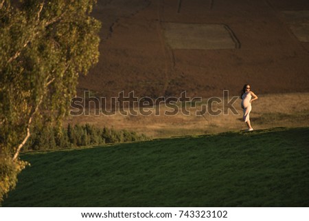 Pregnant Asian woman wearing sunglasses walks between trees with her hands on her belly during sunset in Autumn, 