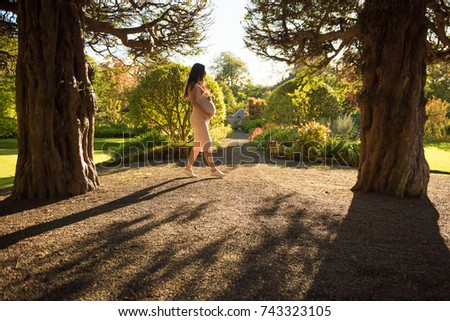 Pregnant Asian woman wearing sunglasses walks between trees with her hands on her belly during sunset in Autumn 
