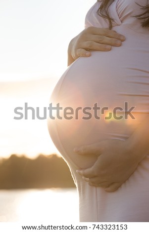 Pregnant Asian woman wearing sunglasses walks between trees with her hands on her belly during sunset in Autumn