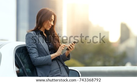 Business Woman Uses Smartphone While Leaning on Her Premium Class Car. Big City with Skyscrapers in the Background. Royalty-Free Stock Photo #743368792