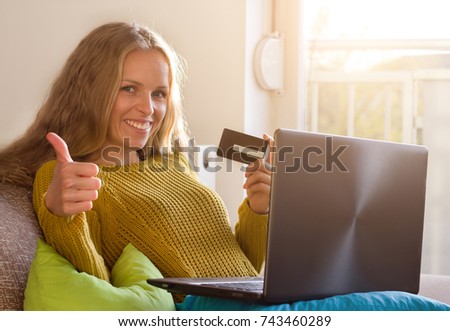 Happy young woman sitting on sofa, holding laptop and credit card and showing thumb up. Online shopping concept