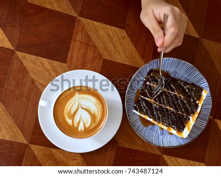 A cup of Cappuccino and a slice of salted Caramel cake from the top with a hand holding a fork.