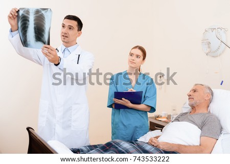 The old man lies on a cot in the medical ward. Next to him is a doctor and a nurse. The doctor looks at the X-ray of the old man. The nurse and the old man also examine the picture.