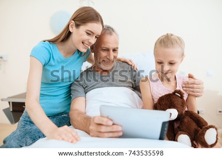 An elderly man lies in a hospital room on a bed. He is seen by a woman with a girl. They are looking at something on the old man's tablet. The girl has a toy in her hands.