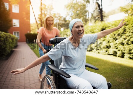 A woman with cancer is sitting in a wheelchair. She walks on the street with her daughter and they fool around. They are fun and they laugh. They walk in the courtyard of the clinic. Royalty-Free Stock Photo #743540647