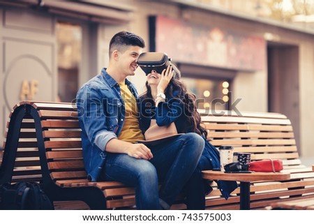 Joyful young beautiful couple boy and girl testing virtual reality 3D video glasses VR headset for game testing in the city impressed by augmented reality on the street with beautiful sun light colors