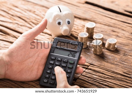 Close-up Of Person Calculating On Calculator With Stack Of Coins And Piggy Bank