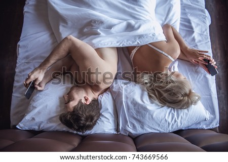 Top view of beautiful young woman and man using their smart phones while lying back to back in bed at home