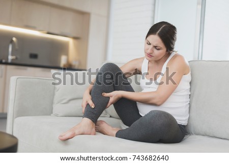 A pregnant woman sits at home on a light sofa. Her legs are numb and she rubs them. Unpleasant for her