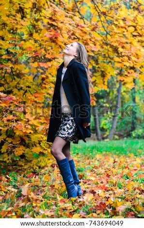 Young girl in classic black coat and blue boots walks in the Park . Autumn Park , green , red and yellow leaves lie on the ground. The girl has long black hair and slim figure .