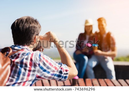 The guy takes pictures of a group of tourists who are sitting in the park on the bench. Behind them is a beautiful landscape. They are posing and smiling.