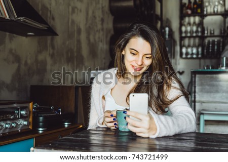 a beautiful brunette girl with a charming smile and expressive brown eyes drinking coffee and using a smartphone, checking social networks, doing photo selfie