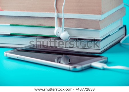Phone with white earphones next to a stack of books on a blue background. The concept of audio books and modern education