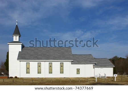 A side view of a three section church outside during the daytime in the winter with a parking area old stained glass windows and a cross on top of the steeple with room for your text.
