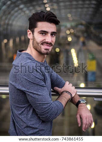 Handsome young man profile shot, indoor, inside big modern building, maybe a brand new train station, looking at camera smiling