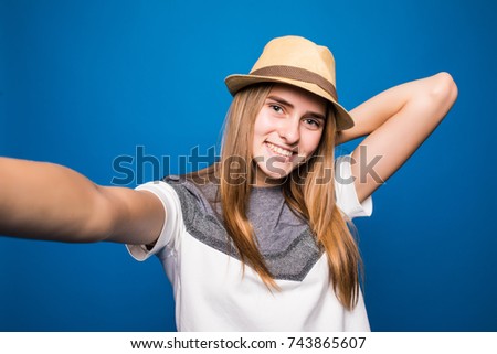 Trendy beautiful hipster girl taking selfie with mobile phone against blue wall.
