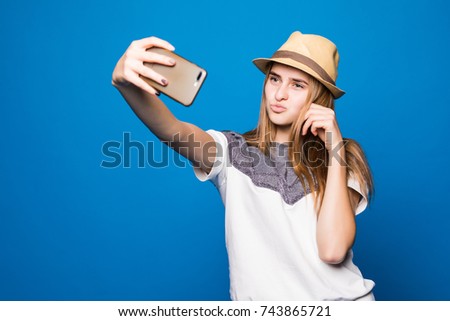 Young l attractive woman on the blue background. She is taking selfie on the camera of her phone, wearing casual summer outfit and a hat