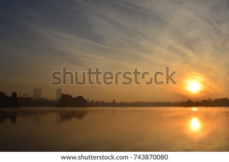 Beautiful morning scene with cloudy sky and sun rising over city lake covered with fog 